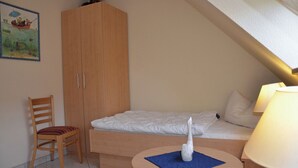 Een kleine, knusse kamer met een enkel bed, een houten garderobe, een stoel en een tafeltje met een decoratief object.