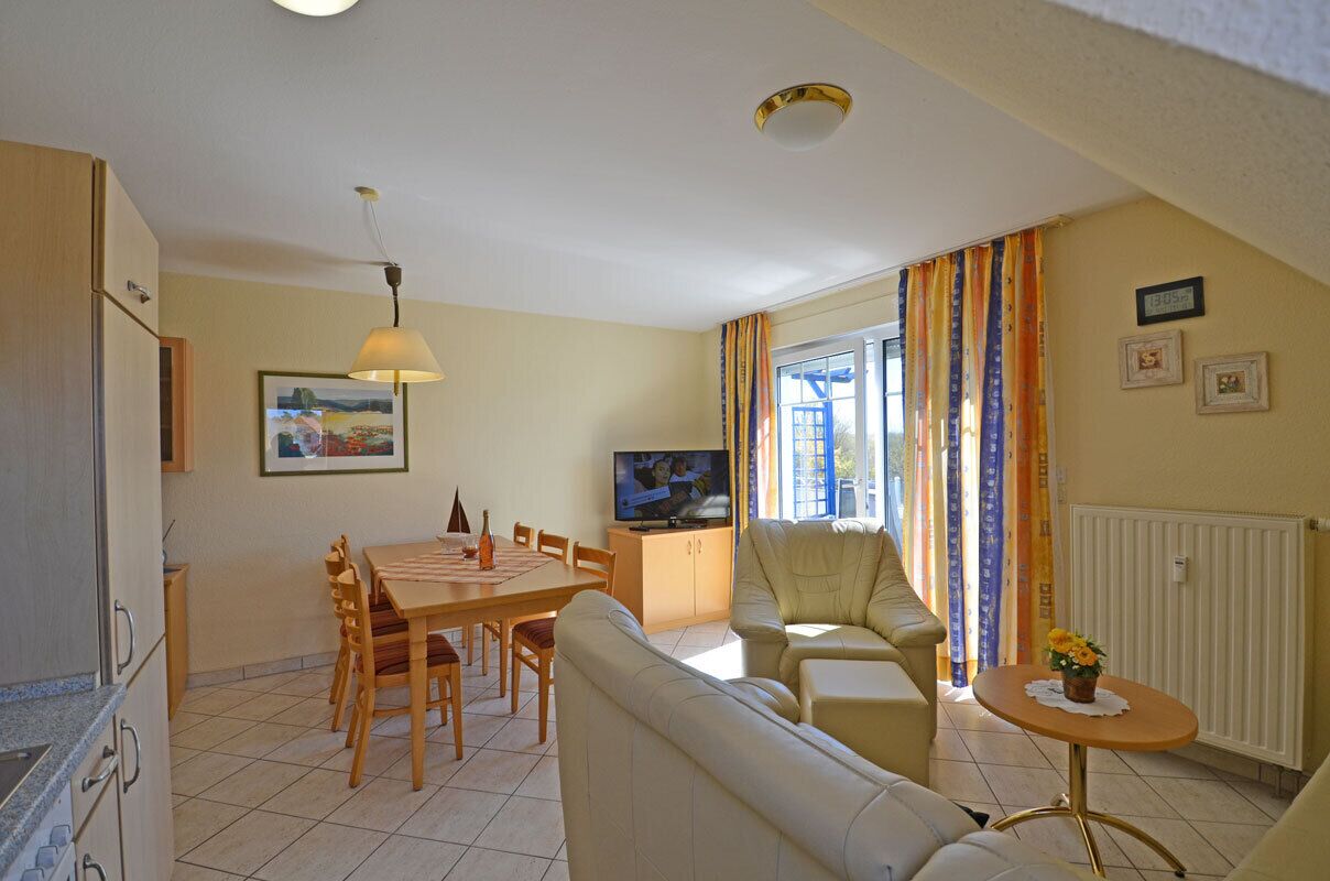 Ein Wohnzimmer mit einer Couch, einem Esstisch mit Stühlen, einem Fernseher und einem Fenster mit Vorhängen.
