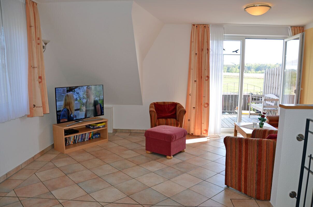 A living room with a television, a red ottoman, striped armchairs, a wooden entertainment unit, and a sliding glass door to a balcony.
