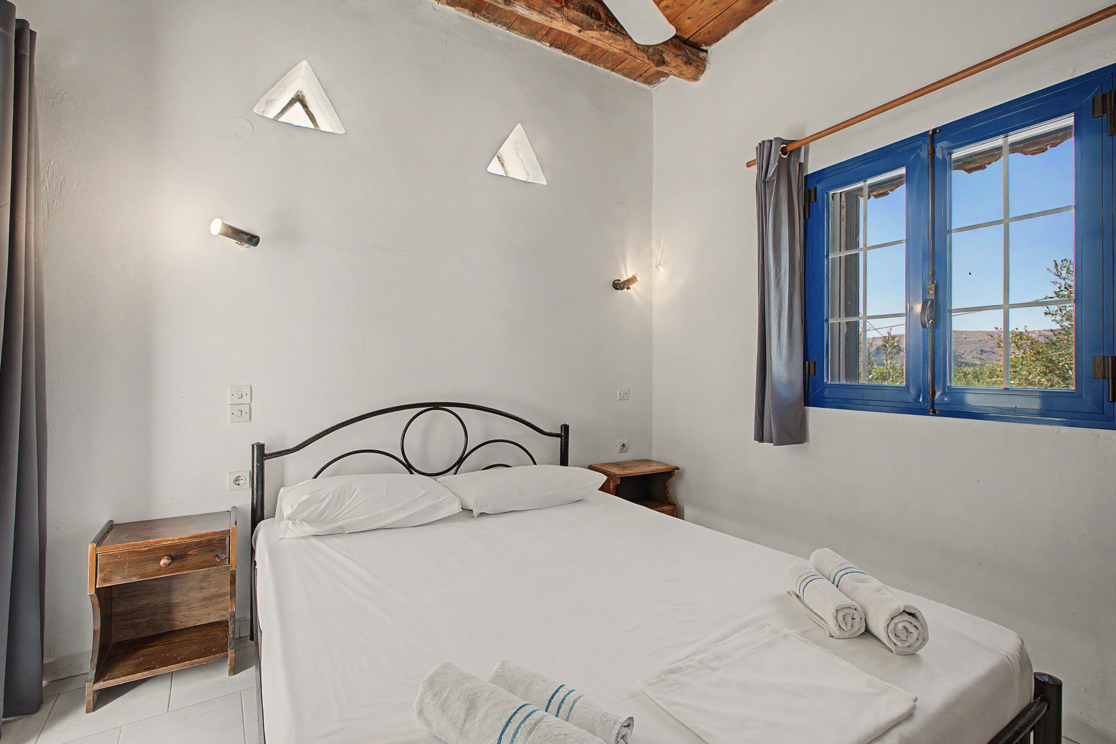 Een slaapkamer met een bed, een houten nachtkastje, een klein tafeltje en een raam met blauwe luiken.
