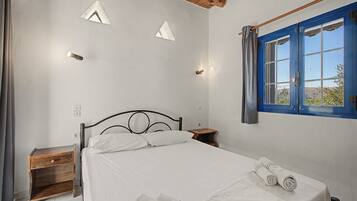 Een slaapkamer met een bed, een houten nachtkastje, een klein tafeltje en een raam met blauwe luiken.