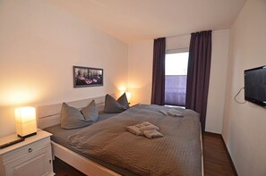 A bedroom with a bed, bedside lamps, a television, a window with curtains, and a picture on the wall. - Just 500 meters from the sandy Baltic Sea beach, Wi-Fi, and a washing machine with a covered patio (Glowe)