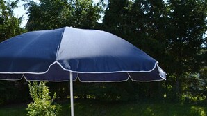 Een terras buiten met een houten tafel en witte stoelen, een blauwe parasol en een vloer van baksteen.