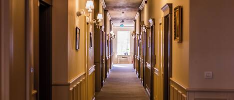 A hotel corridor with doors on both sides, a carpeted floor, and wall-mounted lamps.