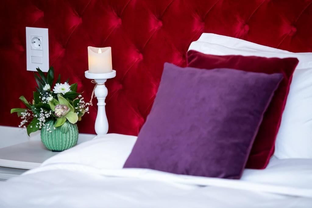 A bed with a red headboard, white bedding, a purple pillow, and a bedside table with a candle and a vase of flowers.
