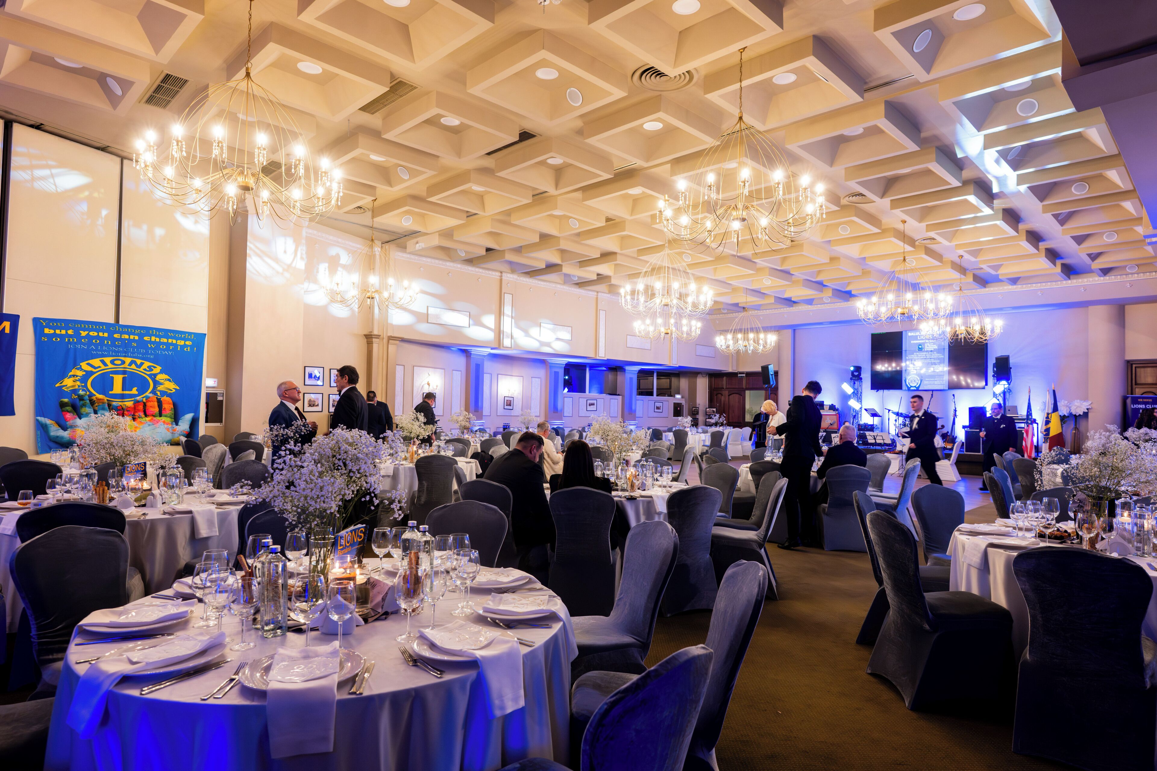 A banquet hall with round tables set for dining, chandeliers, and a stage with musical equipment.