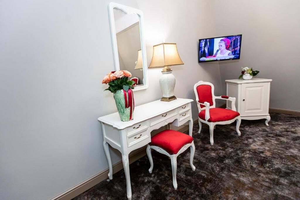 A room with a white vanity, red upholstered chair, a white cabinet, a mirror, a lamp, and a television on the wall.