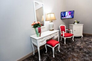 A room with a white vanity, red upholstered chair, a white cabinet, a mirror, a lamp, and a television on the wall.