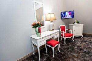 Quarto com toucador branco, cadeira estofada vermelha, armário branco, espelho, luminária e televisão na parede.