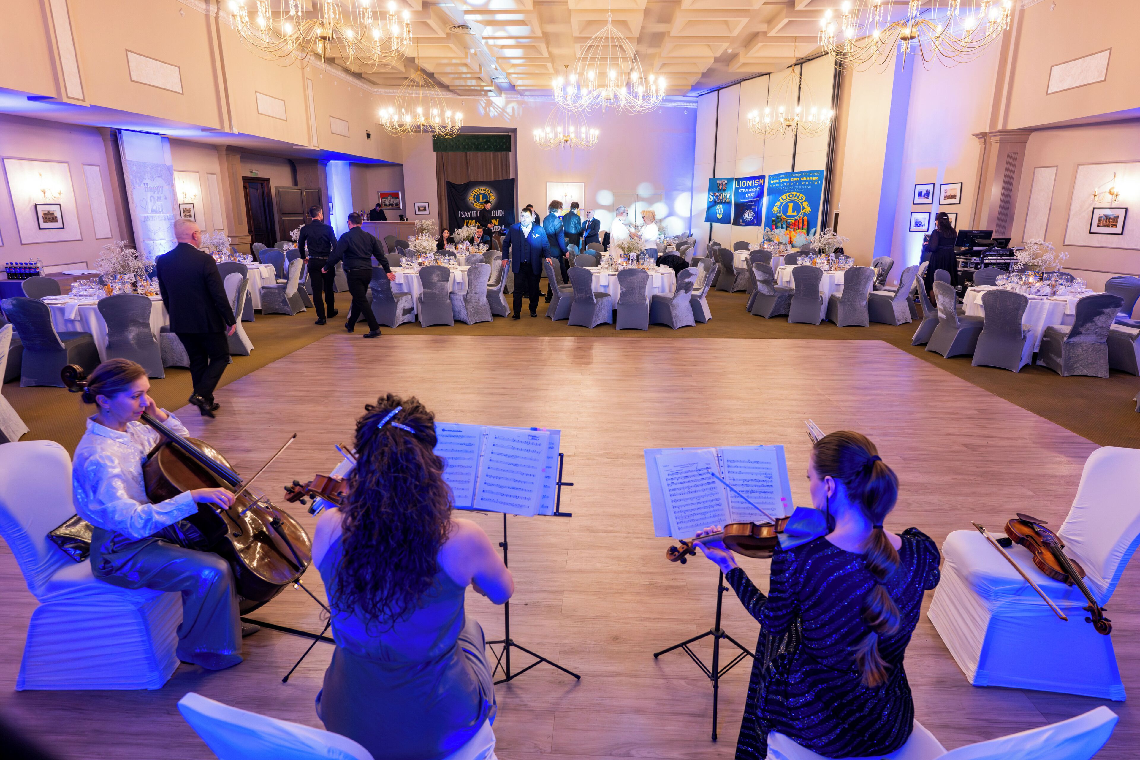 A formal event with seated guests and a live string quartet in a spacious hall with chandeliers and a stage.