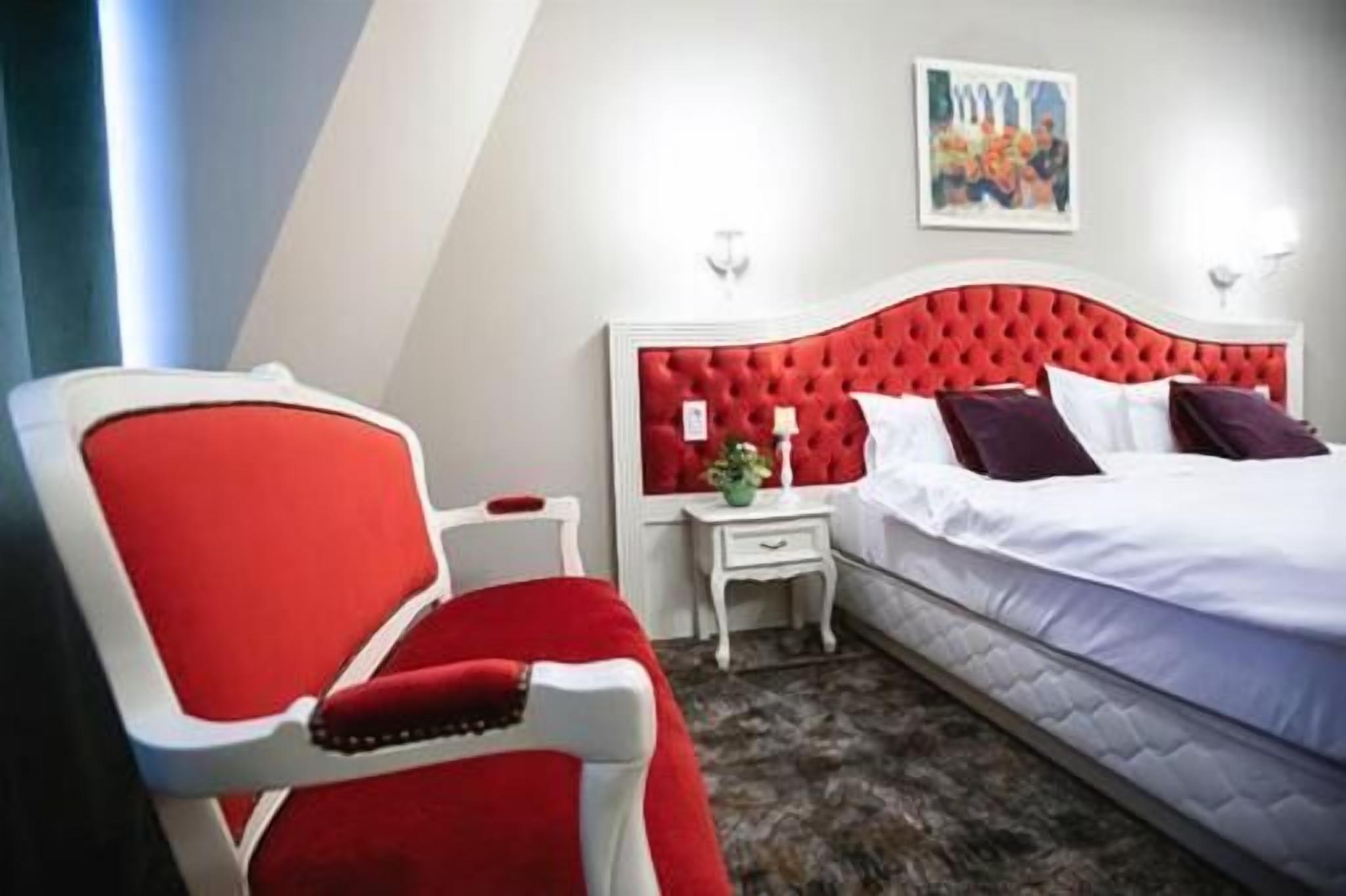 Une chambre d’hôtel avec un fauteuil à revêtement rouge, une table de chevet blanche, une tête de lit rouge et un lit avec du linge de lit blanc.