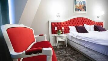 Une chambre d’hôtel avec un fauteuil à revêtement rouge, une table de chevet blanche, une tête de lit rouge et un lit avec du linge de lit blanc.