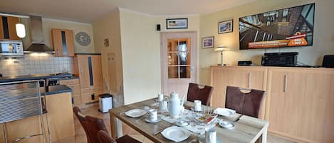 Une cuisine compacte dotée d’appareils électroménagers modernes, un coin repas avec une table en bois pour quatre personnes, et un téléviseur fixé au mur.