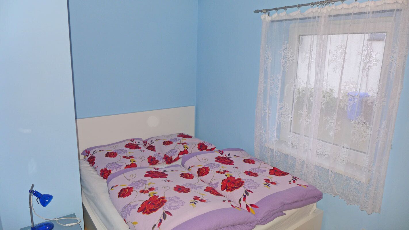 Una cama individual con ropa de cama floral, una lámpara de noche y una ventana con cortinas de encaje.