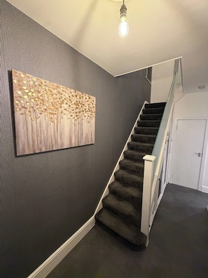 Un escalier recouvert d’une moquette sombre, une ampoule suspendue au plafond et un mur orné d’un motif texturé.