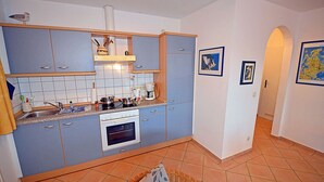 Een kleine keuken met een kookplaat, wastafel en een eethoekje met een rieten stoel.