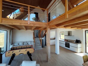 Amplia zona de estar estilo loft con techo de madera, un sofá, una mesa de futbolín y una cocina equipada con electrodomésticos modernos.