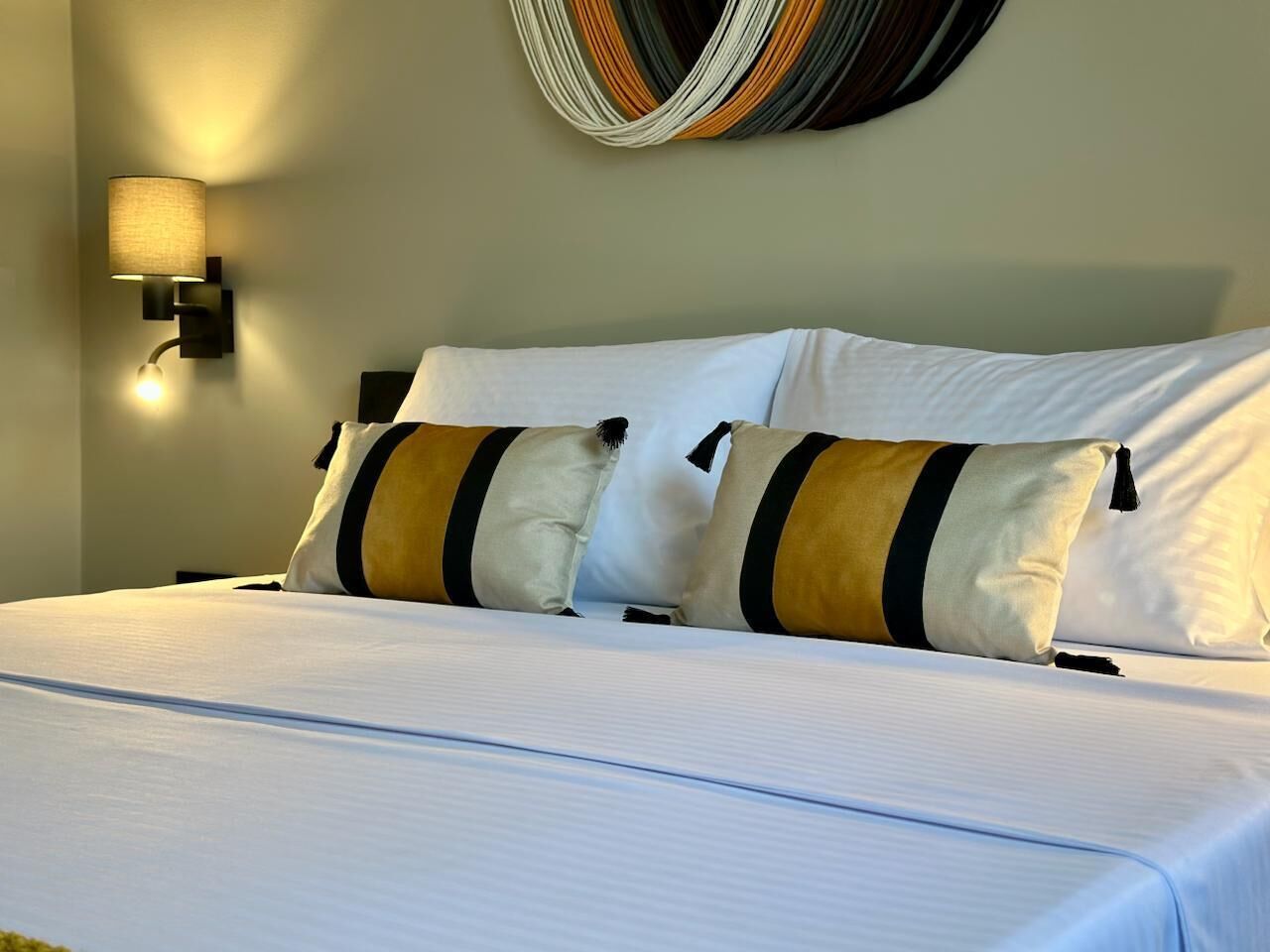 A hotel room with a neatly made bed, two pillows, a wall-mounted light, and a decorative wall hanging.