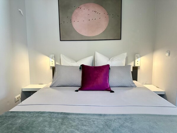 Ein ordentlich bezogenes Bett mit einer weiß-grauen Decke, einem violetten Zierkissen und einer Wandarbeit, die Vögel um einen großen Mond zeigt.