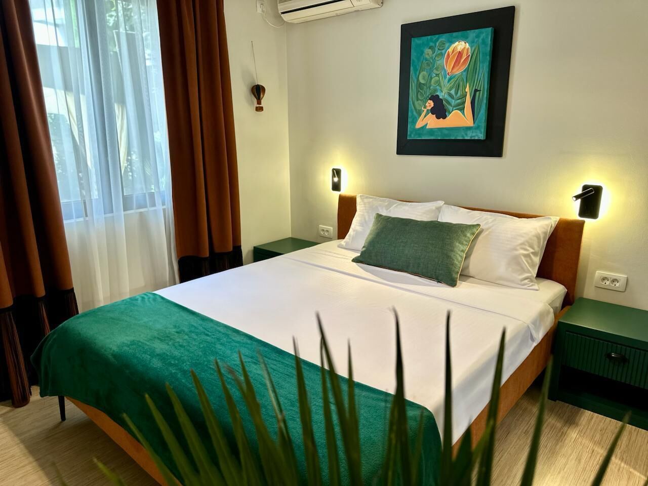 A hotel room with a bed, green curtains, a painting on the wall, and a nightstand with a lamp.