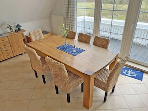 Ein Essbereich mit einem Holztisch, gewobenen Stühlen, einer Vase mit Blumen und Blick auf eine Terrasse durch große Fenster.