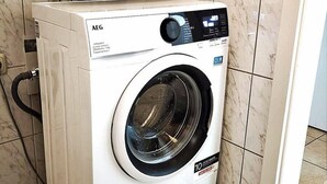 A front-loading white AEG washing machine with a digital control panel, placed in a corner of a room with tiled flooring. - Only 900 meters from the beach, 1st floor with balcony, Internet/Wi-Fi, washing machine (Breege)