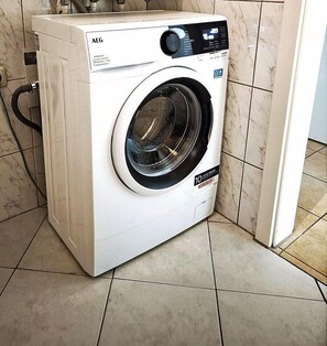 A front-loading white AEG washing machine with a digital control panel, placed in a corner of a room with tiled flooring.