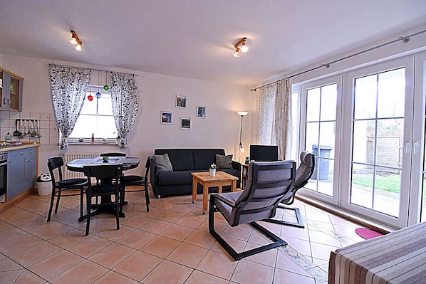 Ein Wohnzimmer mit einer Couch, Sesseln, einem Tisch, einem Fernseher und einem Fenster mit Vorhängen.