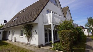 Une maison à deux étages avec un toit en pente, une façade blanche et une véranda entourée d’une clôture.