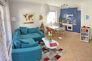 Ein Wohnzimmer mit Sofa, einem Esstisch mit Stühlen, einer Küchenzeile inklusive Schränke und Mikrowelle sowie einem Bücherregal.