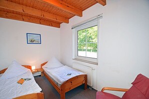 Une chambre avec deux lits en bois, une fenêtre donnant sur un paysage verdoyant, un tableau encadré au mur et une chaise rouge.