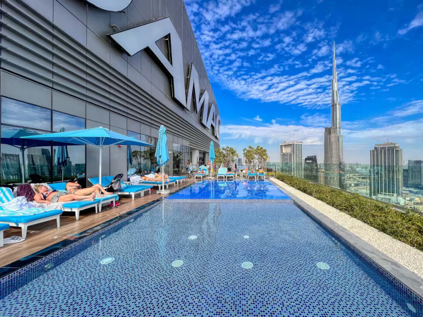 Une piscine sur le toit, agrémentée de chaises longues et de parasols, un grand bâtiment dont la façade porte l’inscription « AM », et une vue sur les toits de la ville avec un gratte-ciel imposant en arrière-plan.