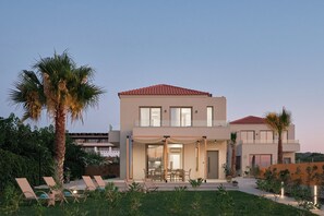 A modern two-story house with a flat roof, large windows, and a covered patio area. - Votsalo Seaside Villa (Kamisiana)