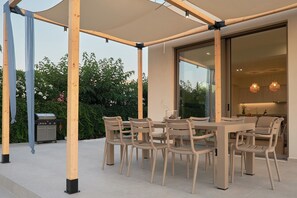 An outdoor patio with a table and chairs, a grill, and a covered area with a view of the garden. - Votsalo Seaside Villa (Kamisiana)