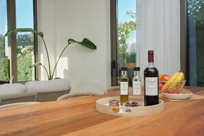 A wooden table with bottles of wine, a bowl of fruit, and a plant in a modern room with large windows. - Votsalo Seaside Villa (Kamisiana)