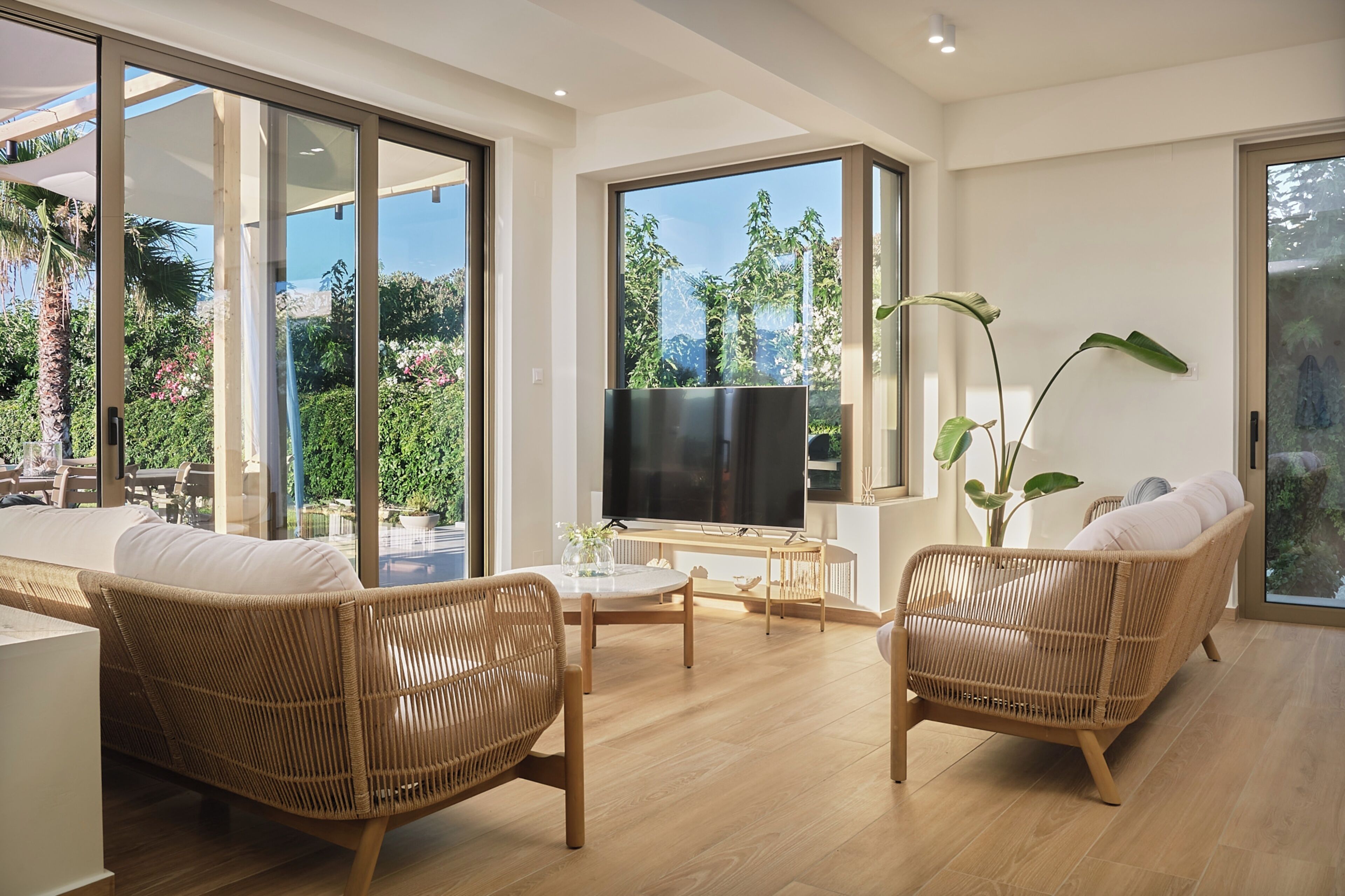Ein modernes Wohnzimmer mit Rattanmöbeln, einem Flachbildfernseher und großen Fenstern mit Blick auf viel Grün.