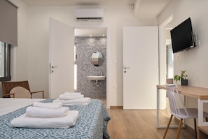 A hotel room with a bed, a desk, a TV, a bathroom with a sink, and a shower. - Votsalo Seaside Villa (Kamisiana)