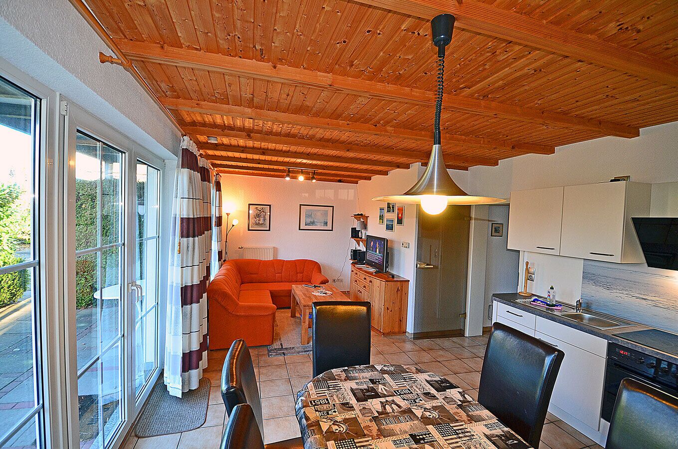 Ein gemütliches Interieur mit Holzbalkendecke, einem Essbereich mit einem gemusterten Tisch und einer Küche mit weißen Schränken.