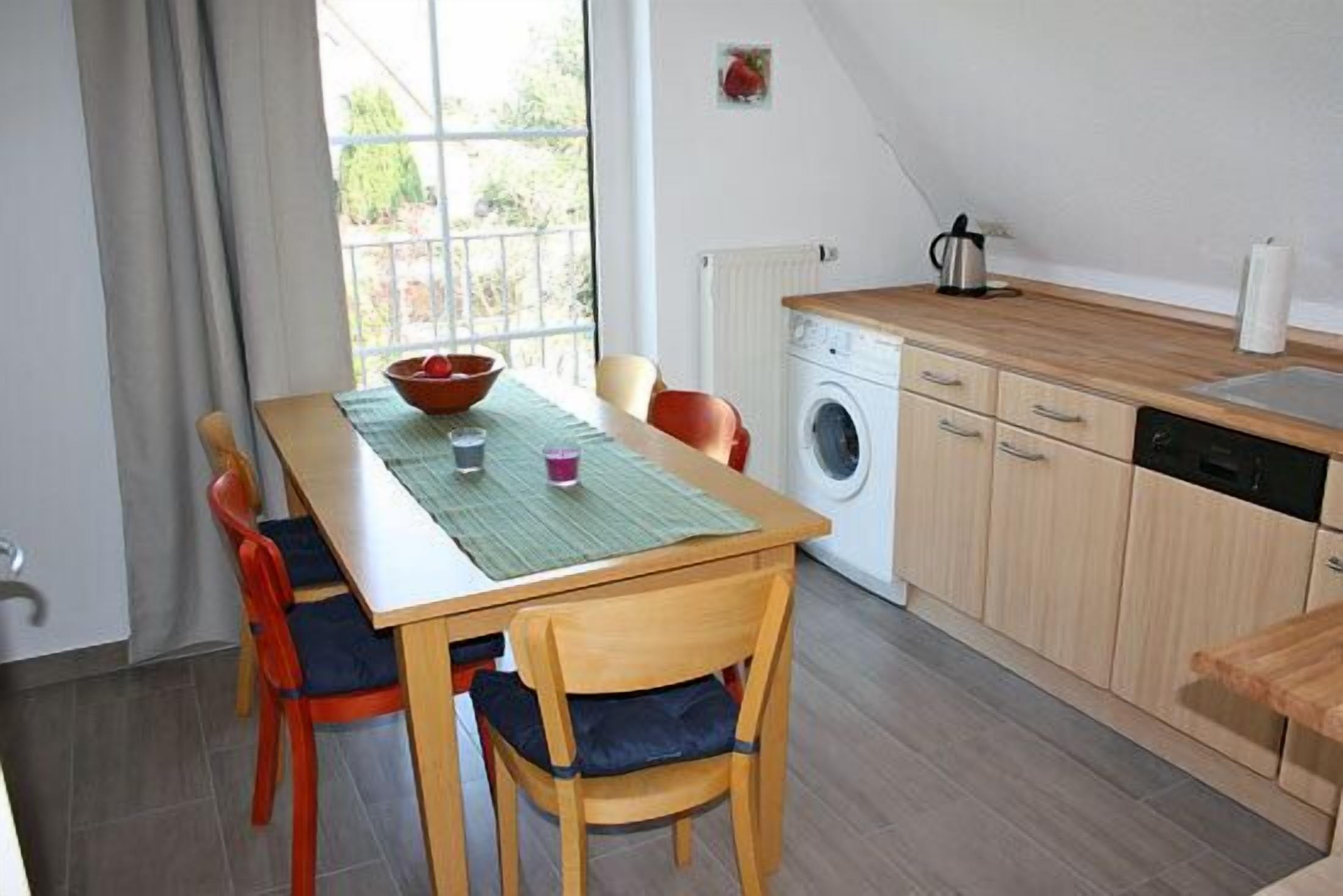A compact kitchen with a washing machine, a dining table with chairs, and a view through a window.