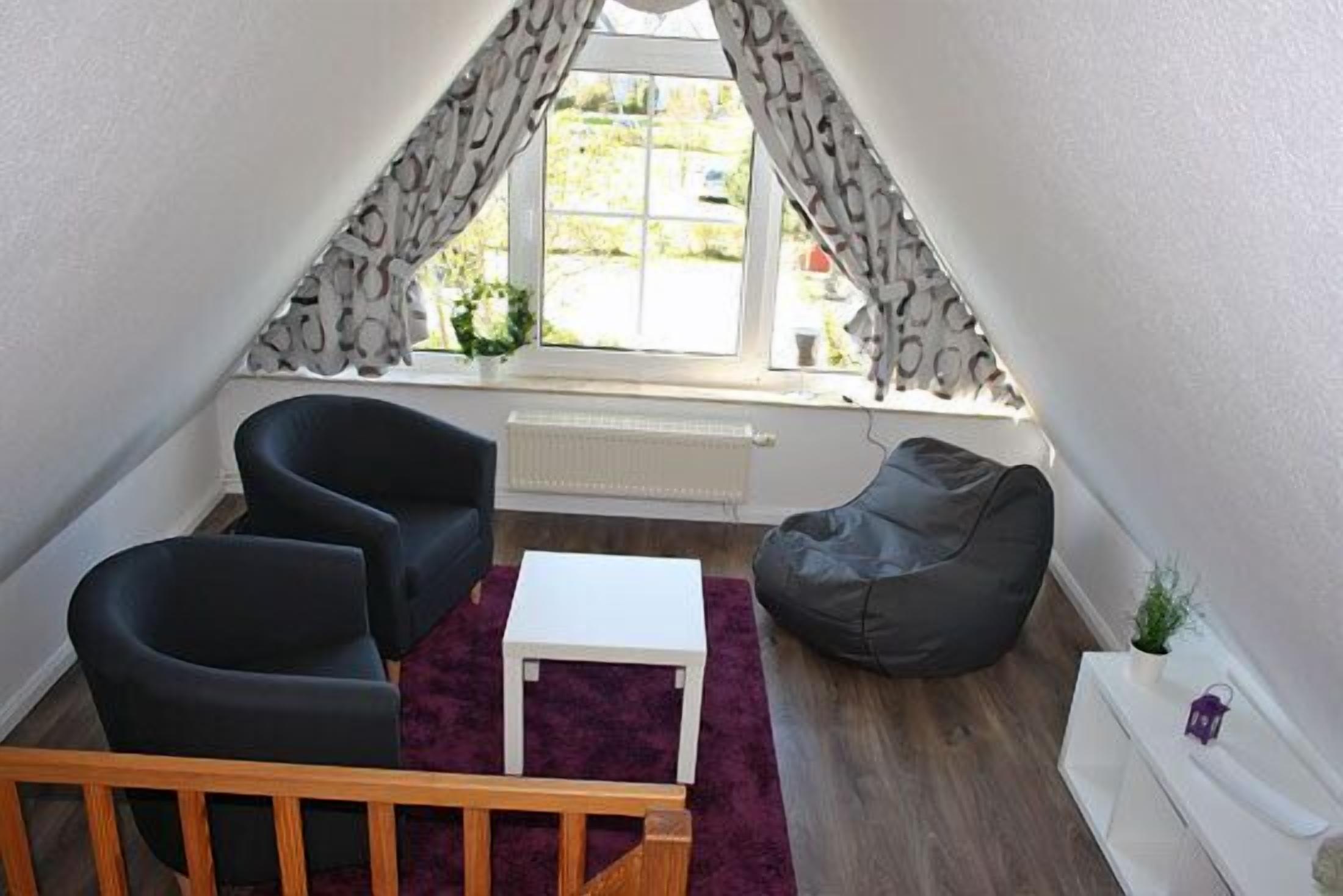A small, triangular attic room with a window, a radiator, a white table, two black chairs, a grey bean bag, and a purple rug.