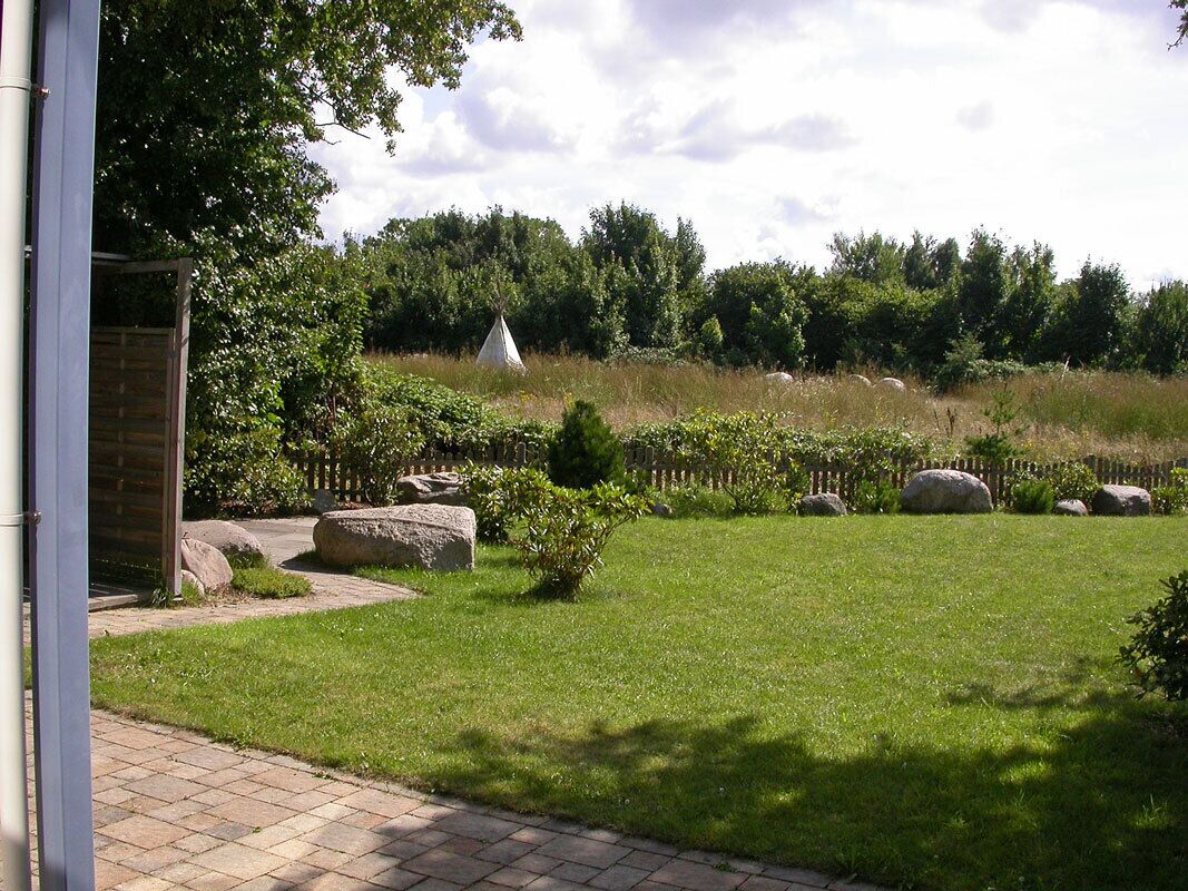 A grassy area with a stone pathway leading to a building entrance, a small garden with rocks, and a white tent-like structure in the background.