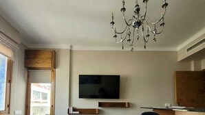 Een moderne woonkamer met een kroonluchter, een flatscreen-tv aan de muur, een houten eettafel met stoelen en een kamerkleed met een patroon op de vloer.