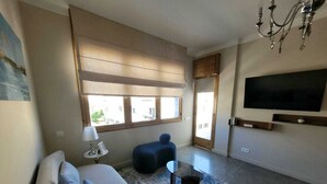Een moderne woonkamer met een bank, een salontafel, een aan de muur bevestigde televisie en een groot raam met zonwering.