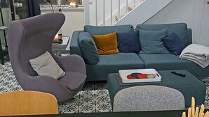 Ein modernes Wohnzimmer mit Sofa, Sessel und Hocker, einem Esstisch mit Speisen und einer Treppe im Hintergrund.