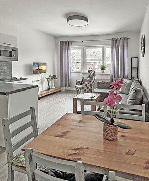 A modern kitchen with a microwave, dining table, and chairs. There is a living area with a sofa, a coffee table, and a TV. A window with curtains is visible. - Vacation rental with a view of the water, sleeps up to 5 people with 2 bedrooms, just 60 meters from the water (Dranske)
