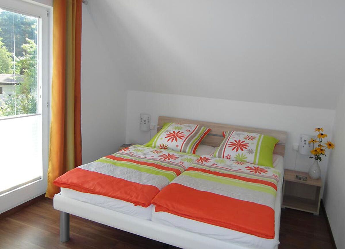 A bed with a white frame and a colorful bedspread, a nightstand with a vase of flowers, and a window with curtains.