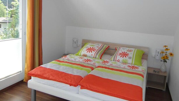 Ein Bett mit weißem Gestell und einer farbenfrohen Tagesdecke, ein Nachttisch mit einer Blumenvase und ein Fenster mit Vorhängen.