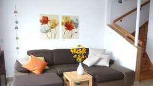 Ein Wohnzimmer mit einem Ecksofa, einem kleinen Holztisch mit Blumen und einer Treppe im Hintergrund.