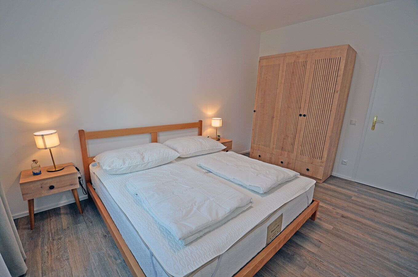 Ein ordentlich bezogenes Bett mit einem hölzernen Kopfteil, zwei Nachttische mit Lampen und ein großer, hölzerner Kleiderschrank.