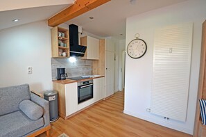 A compact kitchen with a built-in oven, a coffee maker, and a sink. There is a grey sofa and a trash bin in the living area. - Apartment complex in a quiet location, just 80 meters from the water; accommodates up to 2 people (Dranske)